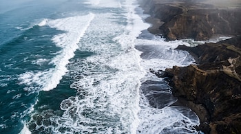 Vista aérea de olas con espuma blanca rompiendo en una playa de arena oscura junto a acantilados rocosos.