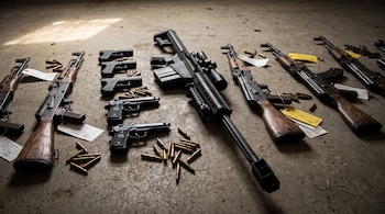 Fusiles de madera, un fusil de gran calibre, pistolas, cartuchos sueltos y tarjetas de evidencia dispuestas sobre un suelo de concreto.