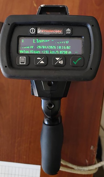 Primer plano de un dispositivo electrónico negro con una pantalla digital iluminada en verde que muestra "PETRNOVIC" y una velocidad de +191 km/h sobre un fondo de madera