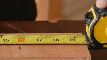 Cinta métrica amarilla con números negros y un dieciséis rojo sobre una pieza de madera, sujetada por una mano