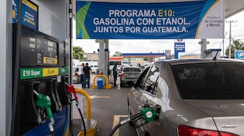 Estación de servicio con surtidores de combustible, mangueras E10 y Super, un automóvil gris, dos hombres y una pancarta azul sobre el programa E10.