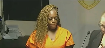Una mujer de cabello largo ondulado lleva un uniforme naranja en una sala de tribunal junto a un micrófono y un sello en la pared
