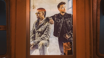 Romeo Santos sentado y Prince Royce de pie a su lado, ambos vistiendo casacas de mezclilla dentro del marco de una ventana