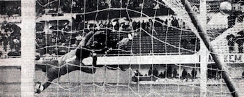 Fotografía en blanco y negro de un portero de fútbol lanzándose para atajar un balón en un arco con red, con espectadores en las gradas
