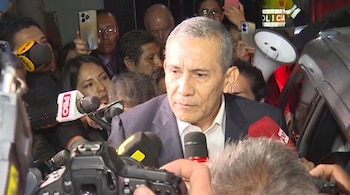 Hombre canoso en traje rodeado por micrófonos de prensa, cámaras de fotos, teléfonos celulares y otras personas. Policía visible a la derecha