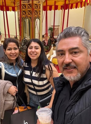 Primer plano de un hombre con barba gris tomando una selfi con dos mujeres sonrientes; detrás, un carrusel con caballos y decoración dorada, y bolsas de compra
