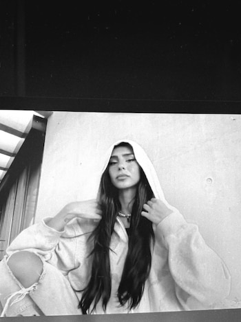 Juana Tinelli en blanco y negro, sentada, con sudadera con capucha, jeans rotos y collar. Su largo cabello oscuro cae. Mira a cámara