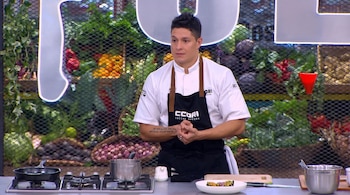 Un cocinero con chaqueta blanca y delantal negro permanece de pie ante una encimera con fogones, ollas y un plato preparado.