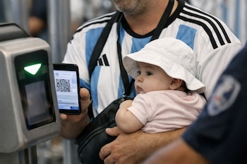 Hombre con camiseta argentina, bebé con sombrero blanco, teléfono móvil con código QR, molinete con luz verde y parte de un guardia de seguridad.