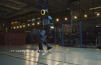 Robot humanoide azul y negro con cabeza brillante amarilla, realizando una finta en suelo claro con conos amarillos y escalera de agilidad en un laboratorio