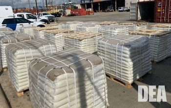 Pallets con bultos rectangulares envueltos en plástico y atados con cuerdas. Al fondo hay vehículos, contenedores de envío y edificios portuarios. Logo de la DEA
