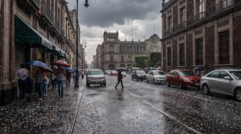 Calle con edificios, personas con paraguas, vehículos circulando y estacionados, granizo sobre el asfalto mojado y lluvia cayendo bajo cielo nublado.