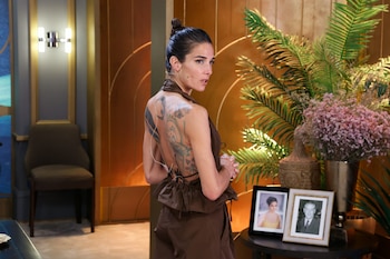 Juana Viale de espaldas con una prenda marrón de espalda abierta que muestra sus tatuajes, junto a arreglos florales y fotografías