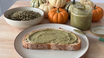 Una rebanada de pan con crema verde sobre un plato, un cuenco con semillas, un frasco transparente y varias calabazas pequeñas al fondo.