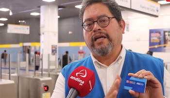 Hombre con barba y gafas, con chaleco azul, sostiene una tarjeta de transporte azul y habla a un micrófono rojo con el logo de Andina en una estación de metro