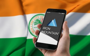 Mano sostiene un teléfono móvil con el logo de Iron Mountain en la pantalla. Al fondo, una bandera con franjas naranja, blanca y verde, y un sello de ciudad borroso