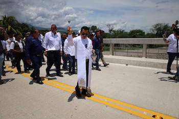 Un sacerdote con vestimenta blanca bendice con un aspersorio sobre una carretera, flanqueado por hombres. Al fondo, la barandilla de un puente, montañas y árboles