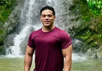 Un hombre de cabello oscuro y camiseta granate sonríe a la cámara, de pie en primer plano frente a una cascada de agua y follaje verde