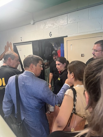 Grupo de personas, incluyendo un hombre de traje azul y varias personas con camisas con la insignia 'DI', paradas en un pasillo con puertas numeradas '6' y '7'