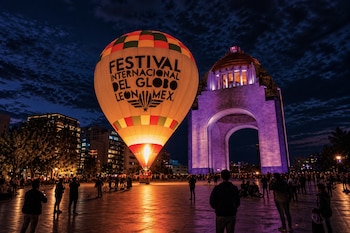 Un globo aerostático encendido permanece en una plaza nocturna junto a un monumento iluminado de morado y rodeado por personas.