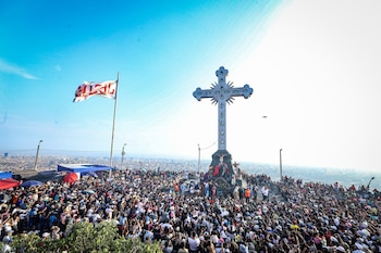 Vista aérea de una gran multitud en el Cerro San Cristóbal, con una imponente cruz blanca y una representación del Vía Crucis. El cielo azul y la ciudad de Lima al fondo
