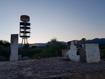 Vista de una torre metálica deteriorada con la palabra 'ORO' en la parte superior, edificaciones en ruinas, escombros y montañas al fondo bajo un cielo despejado