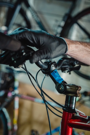 Primer plano de unas manos enguantadas en negro sujetando el manillar de una bicicleta roja. Se observa parte del cuadro, los cables y una cinta azul