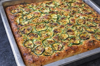 Vista cenital de una focaccia de calabacín entera en una bandeja para hornear, cubierta con rodajas de calabacín, perejil y sal.