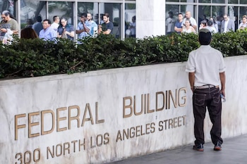 Un hombre de espaldas; muro con letras grabadas 'FEDERAL BUILDING 300 NORTH LOS ANGELES STREET'; arbustos verdes; personas frente a un edificio de cristal