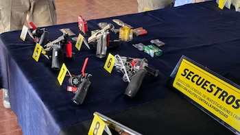 Varias armas de fuego, cajas de balas, cartuchos y marcadores de evidencia amarillos numerados sobre una mesa cubierta con mantel oscuro