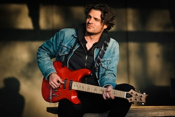 Mark Lettieri, guitarrista de Snarky Puppy, sentado y tocando una guitarra eléctrica roja con una correa estampada. Viste chaqueta vaquera y sudadera oscura