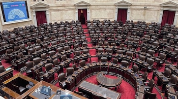 Recinto legislativo con bancas de cuero marrones vacías, escritorios de madera, alfombra roja, paredes claras, cortinas rojas, pantallas electrónicas y un micrófono.