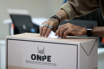 Primer plano de manos de una persona depositando una papeleta blanca en una urna electoral de cartón con el logo de ONPE en un centro de votación