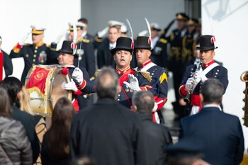 Un director de banda militar con uniforme azul y rojo, sombrero de copa y guantes blancos, gesticula con las manos frente a otros músicos y una multitud