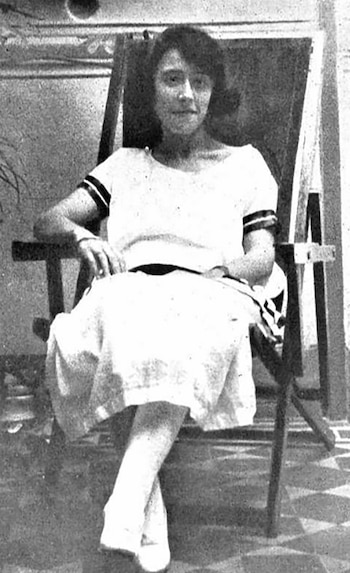 Fotografía en blanco y negro de una mujer joven sentada en una silla de lona, mirando a la cámara. Viste una blusa clara con rayas oscuras en las mangas