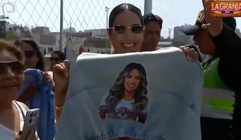 Pamela López sonriente, con gafas de sol, sostiene una camiseta con su imagen impresa. La rodean personas y se ve una valla de fondo bajo el sol