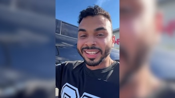 Primer plano de un hombre sonriente con barba y cabello oscuro, camiseta negra, parado frente a una camioneta oscura bajo un cielo azul claro