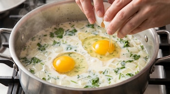Primer plano de manos rompiendo un huevo en una olla grande con leche, cebolla picada y hierbas verdes, donde ya hay dos huevos más cocinándose.