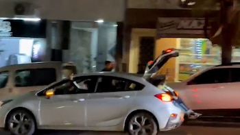 Un coche hatchback blanco con el maletero abierto visible desde la parte trasera derecha, con otros coches y edificios iluminados en el fondo