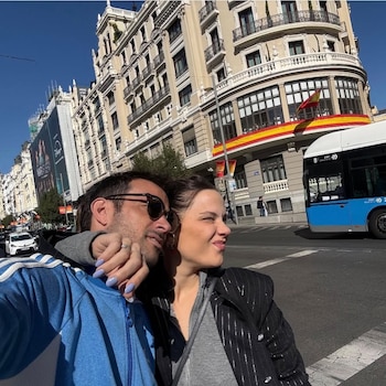 Un hombre y una mujer posan para una selfie en una calle urbana; se ve un autobús azul, edificios históricos y una bandera española en el fondo
