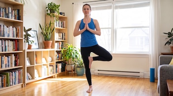 Una mujer con top azul y mallas negras realiza una postura de yoga. Hay estanterías, plantas, una ventana grande y un sofá.