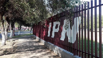 Vista de una valla metálica de color rojo oscuro con las letras "IPN" en blanco, junto a una acera y árboles con troncos pintados de blanco