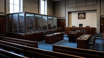 Bancos de madera, mesas con micrófonos, una estructura acristalada y el escudo real británico en la pared de una sala de tribunal.