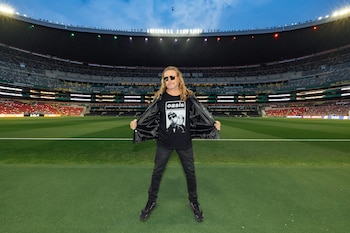 Hombre con cabello rubio largo, gafas de sol, chaqueta y camiseta negra con logo 'Oasis' posa en césped de un estadio con gradas vacías y pantallas