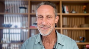 Primer plano de un hombre de mediana edad, sonriente, con cabello gris y barba, camisa azul claro y un collar, frente a una estantería de madera desenfocada