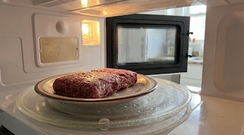 Bloque de carne picada cruda en un plato marrón sobre el plato giratorio de un microondas blanco con la puerta abierta y la luz interior encendida.