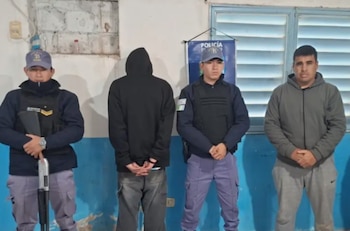 Cuatro hombres parados, dos policías uniformados con gorras y chalecos, uno sostiene una escopeta. Dos hombres vestidos de civil, uno de espaldas, otro de frente. Banderín con texto Policía