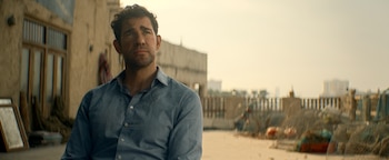 Un hombre con camisa azul, barba ligera y una marca en la frente mira hacia arriba con expresión seria, con edificios de adobe al fondo