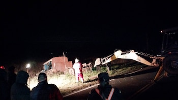 Vista nocturna de un accidente en la carretera, mostrando un camión volcado, una retroexcavadora amarilla y negra, y varias personas