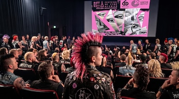 Personas con peinados de cresta y chaquetas de cuero de espalda a la cámara contemplan una pantalla de cine proyectada.